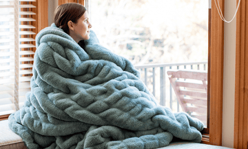 Woman wrapped in a Glacier Glide Chic minky blanket from Minky Designs, sitting by a bright window and showcasing the plush, braided texture and cozy feel of the Chic collection.